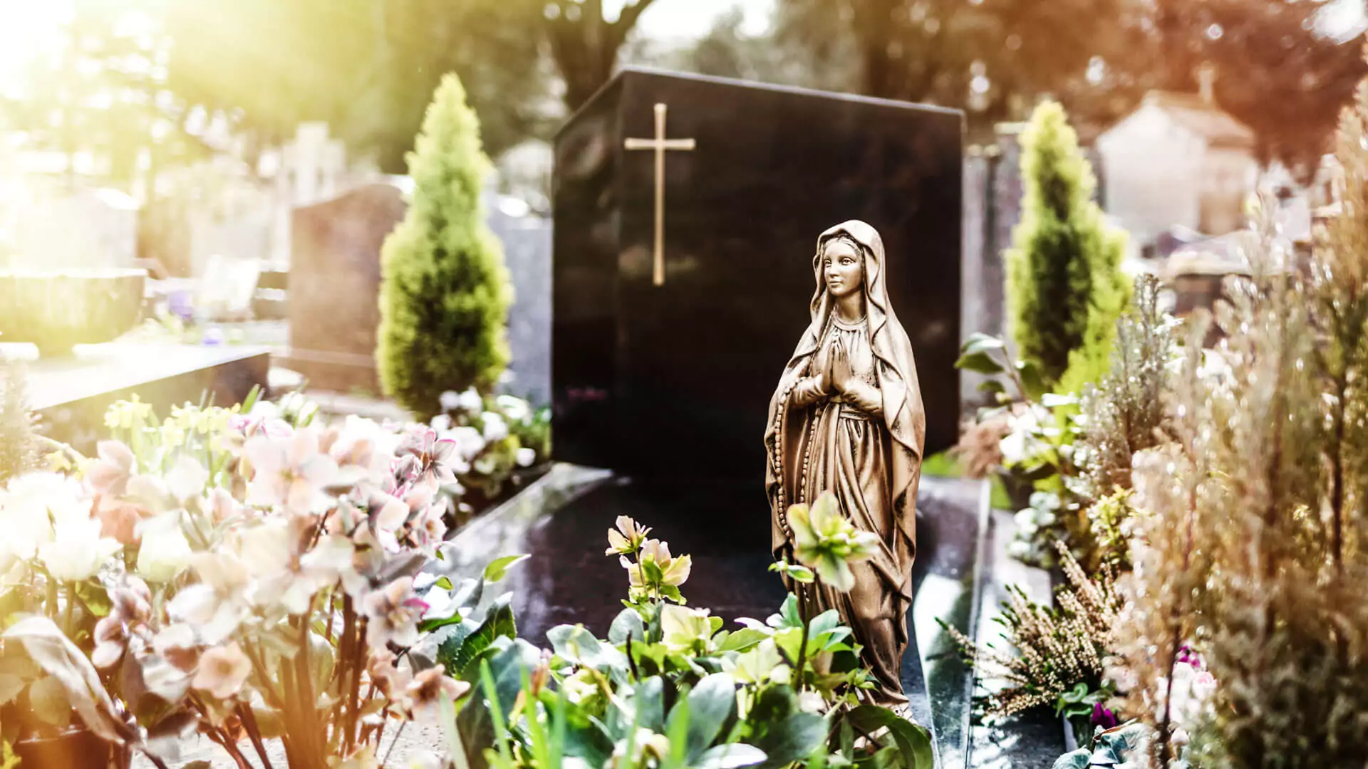 Grabstein mit goldenem Kreuz und davor steht eine Figur der Jungfrau Maria mit Blumenschmuck rund um das Grab auf einem Friedhof.